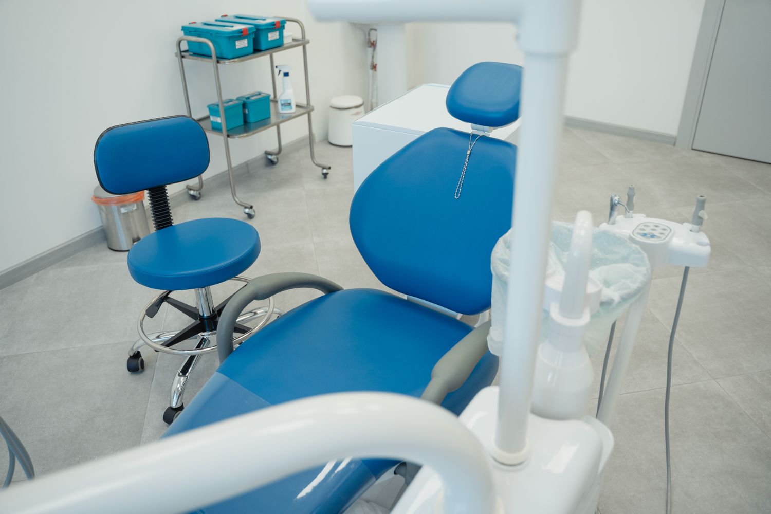 Blue dental chair and stool beside a steel trolley.