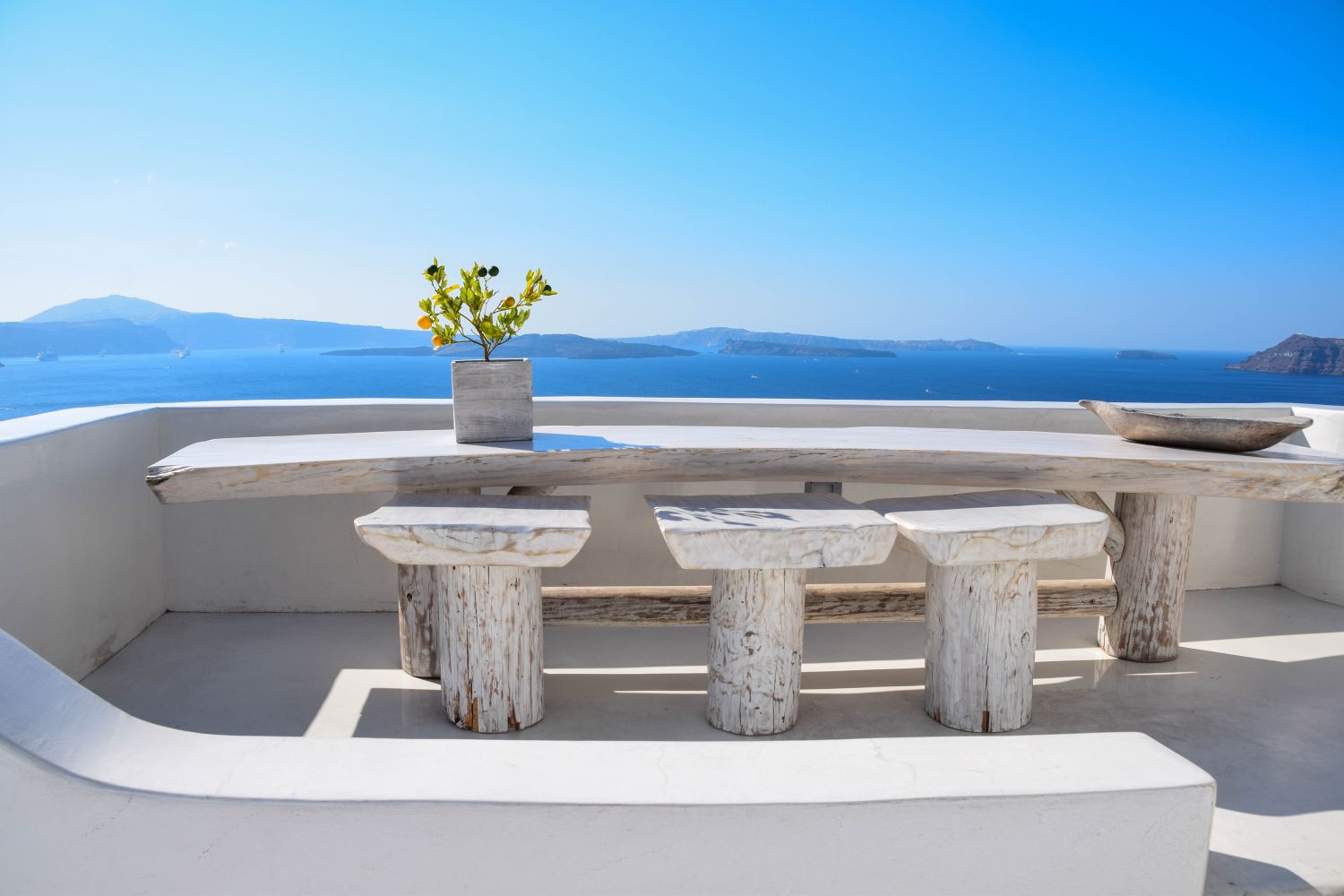 A whitewashed holiday terrace with a table and stools, the sea and islands beyond